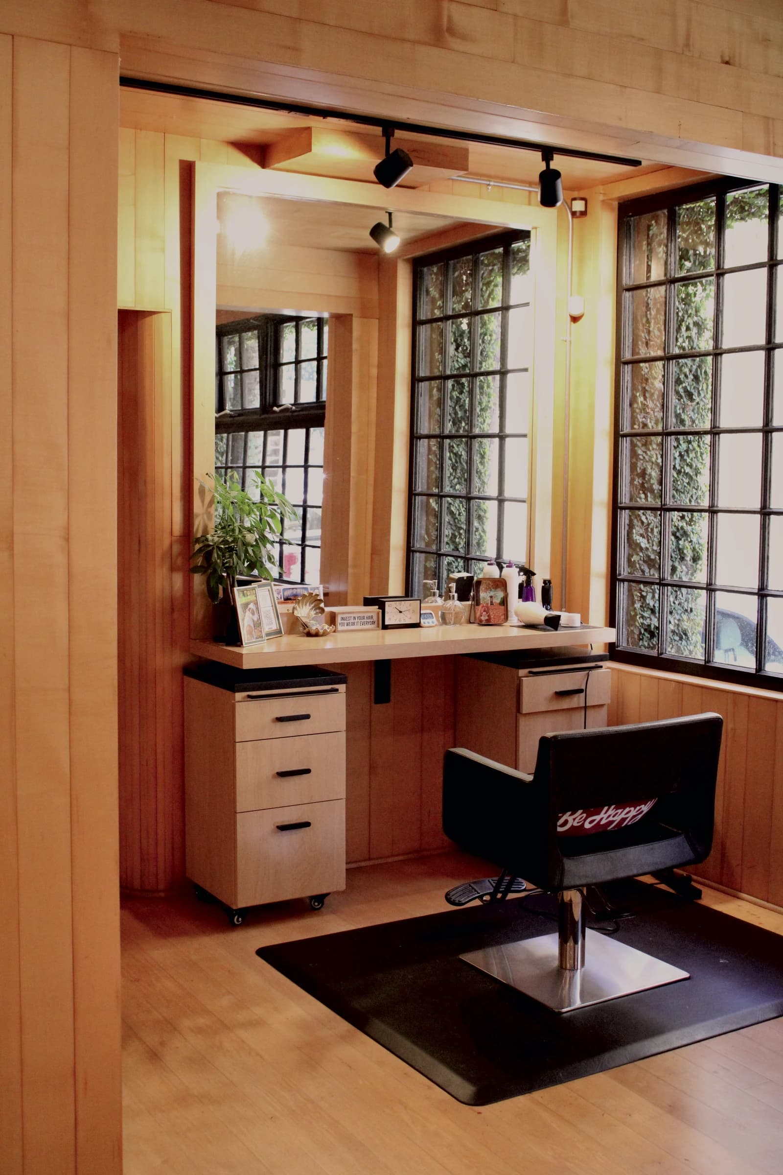 A styling station in a maple alcove: a black chair on a floor mat, a mirror over a counter with drawers, tall black-framed windows