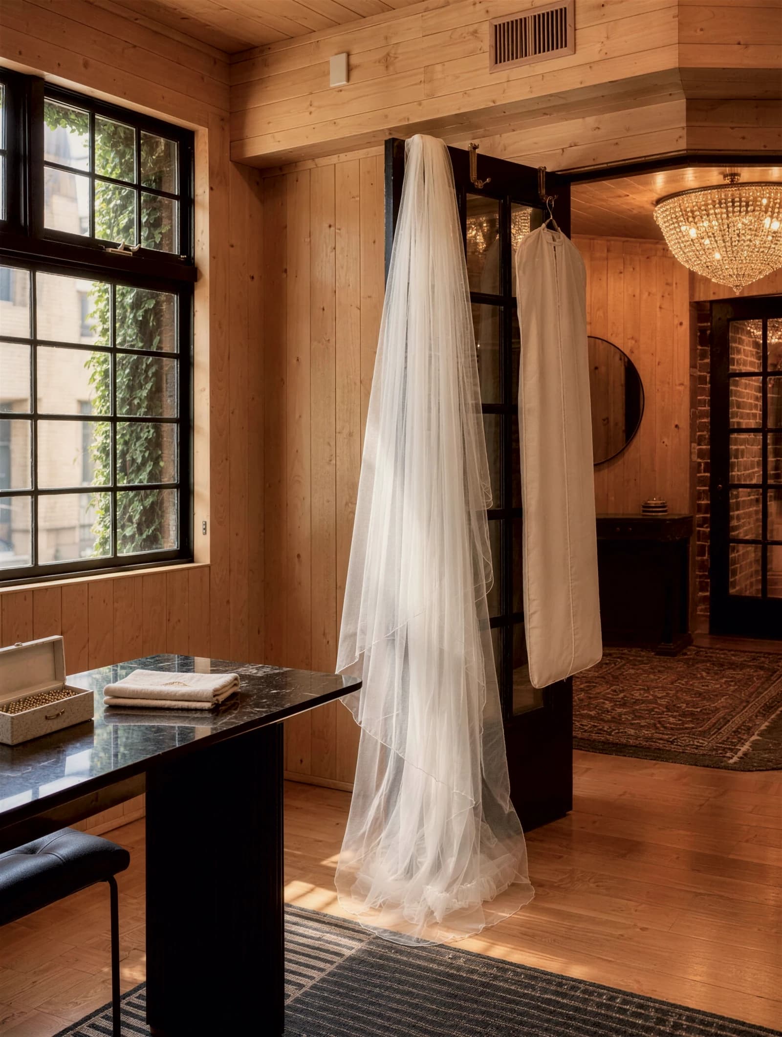 Rendering of a bridal veil and a garment bag hanging on an open black-framed door in a maple-paneled room, a crystal chandelier beyond