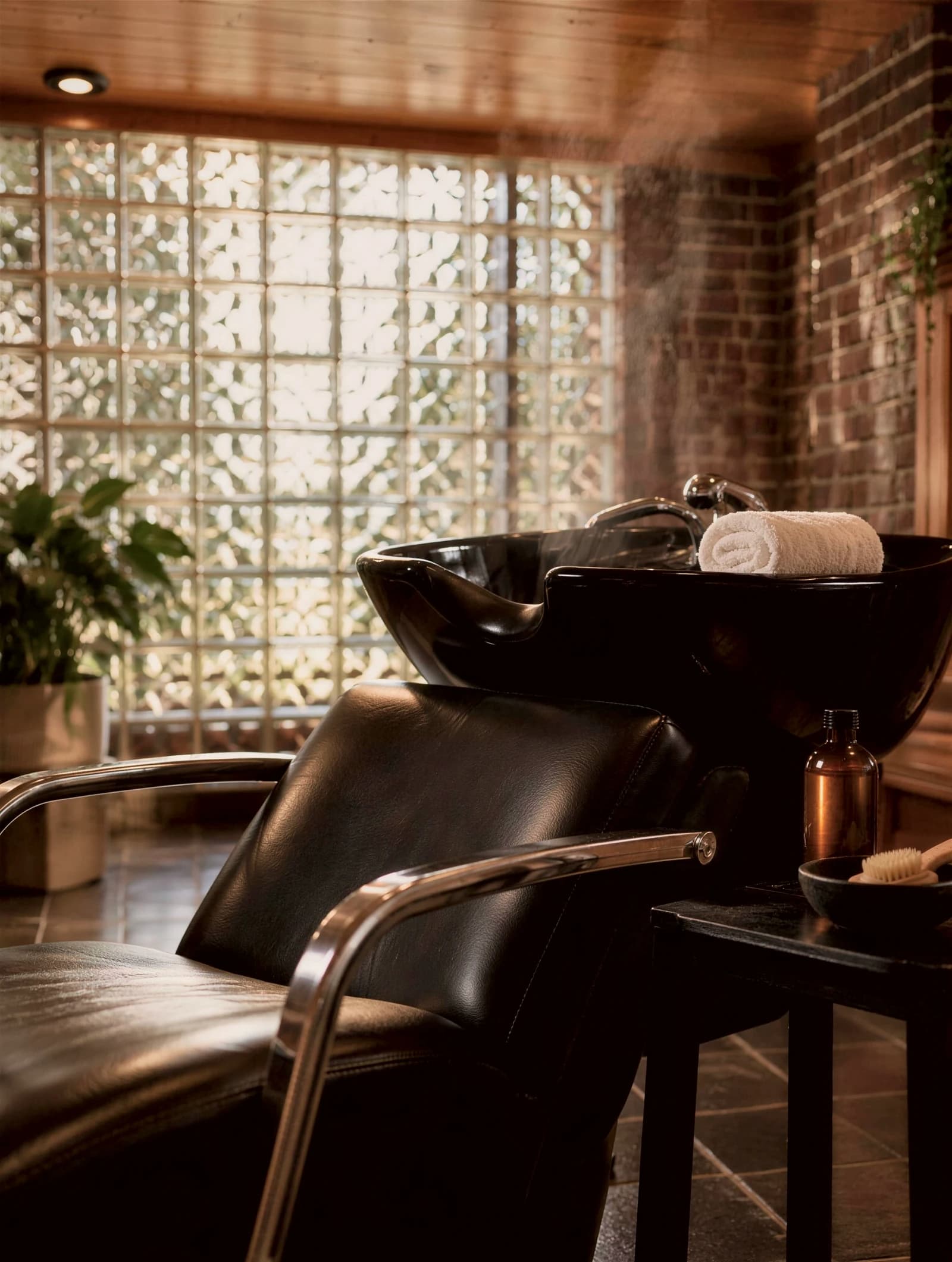 Rendering of a black leather shampoo chair and basin with a rolled white towel, in front of a glass-block wall