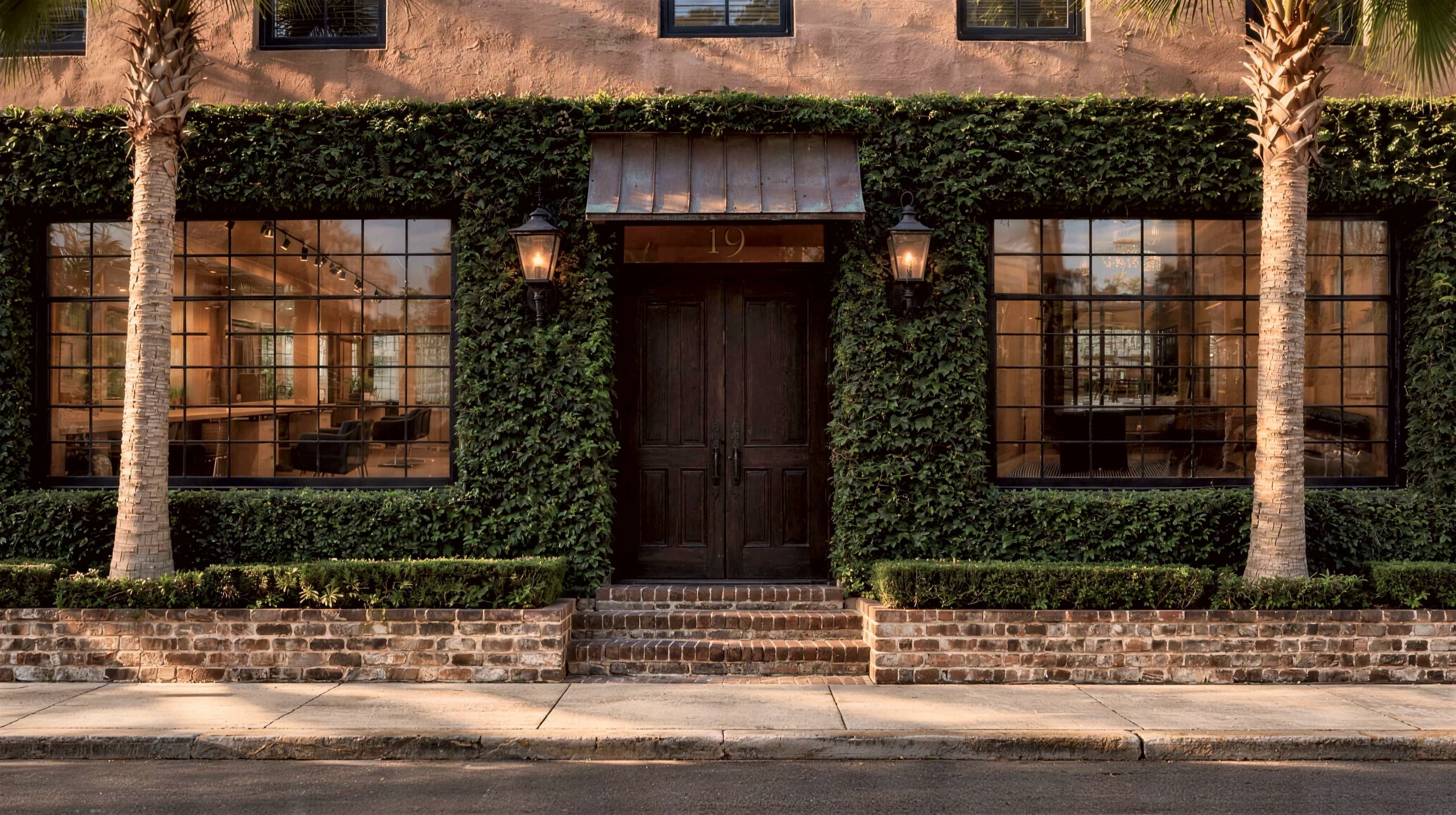 Composite rendering of the salon's entrance: a dark double door under a copper awning marked 19, two lit lanterns, ivy-covered walls with a wide window on each side, palms and low brick planters along the sidewalk