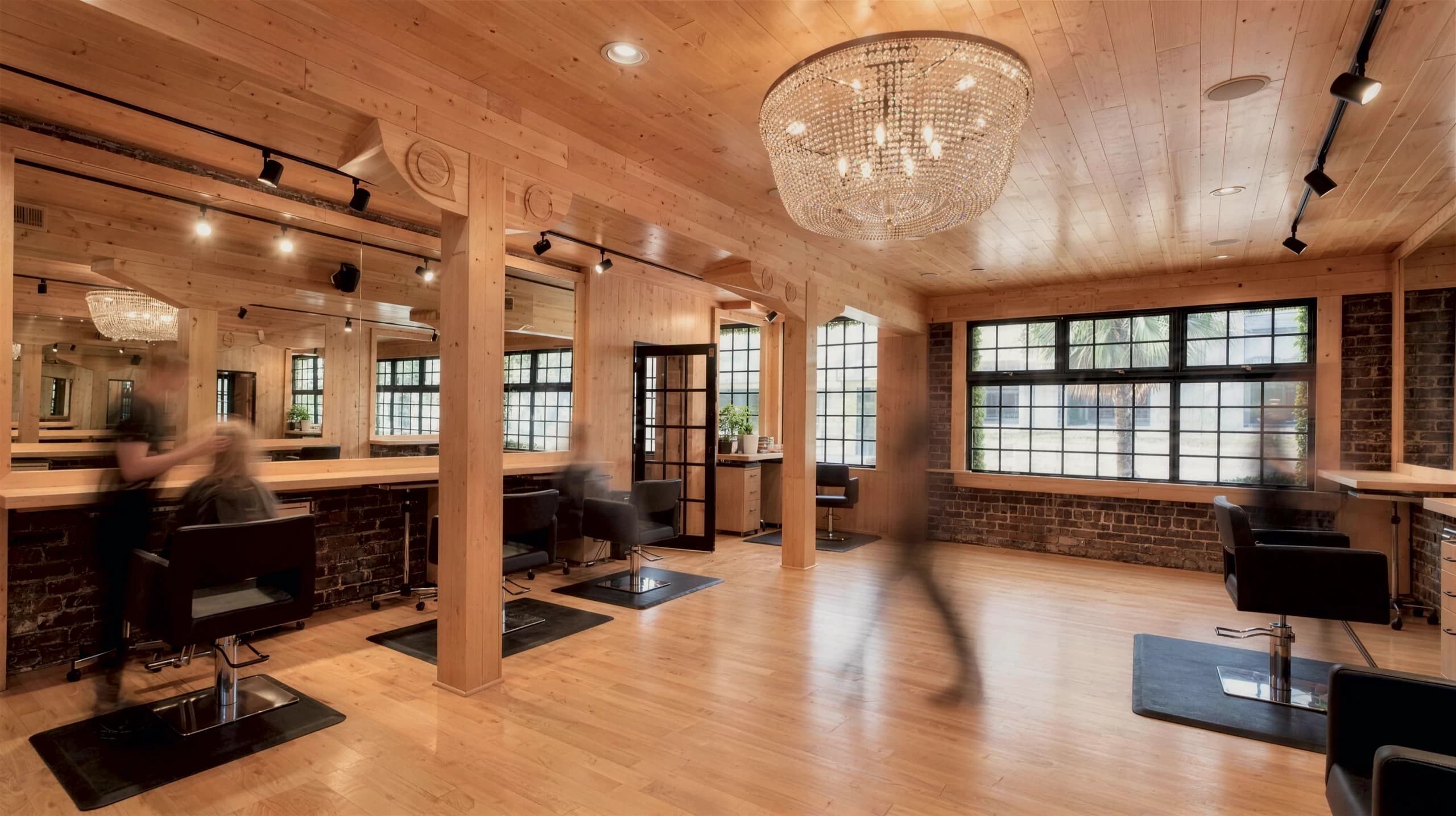 Long-exposure rendering of the salon floor: black styling chairs along maple counters and mirrors, maple columns, a crystal chandelier, a wide black-framed window over a brick wall, and blurred passing figures